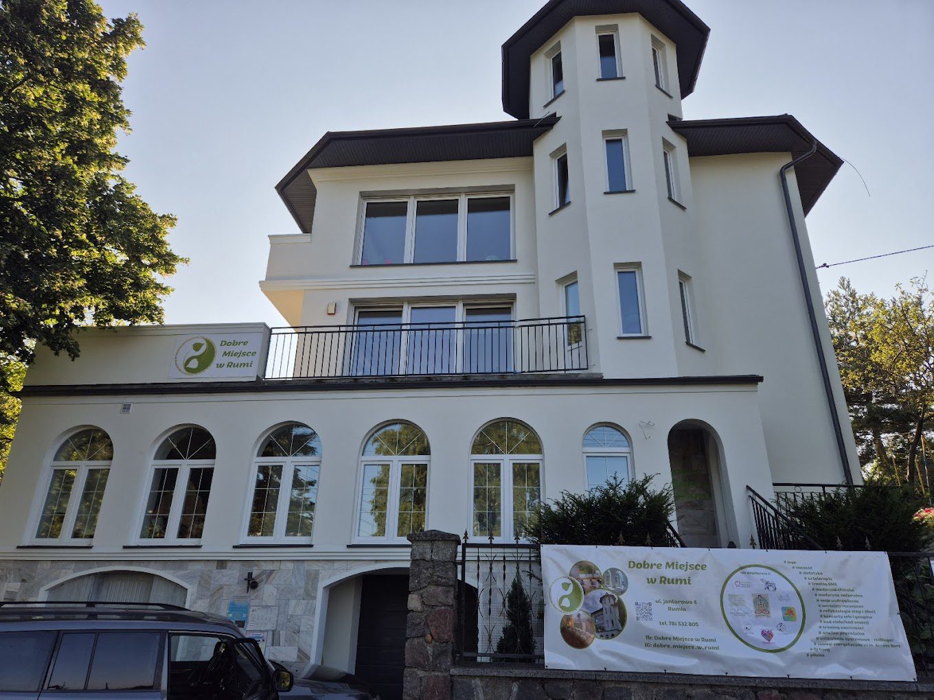 biały dom z wieżyczką w otoczeniu lasu, na ogrodzeniu reklama Dobrego Miejsca w Rumi
